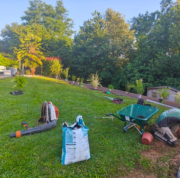 Jardinage et plantation : aménagement paysager avec terreau. Aménagement paysager sur pelouse en pente. Brouette, sac de terreau, souffleur et plantes alignées pour la plantation.